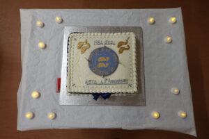 A square cake with 1984 - 2024 HoTS 40th Anniversary written on it in icing with the Tetbury Dolphins, top left and the tetbury coat of arms in the centre